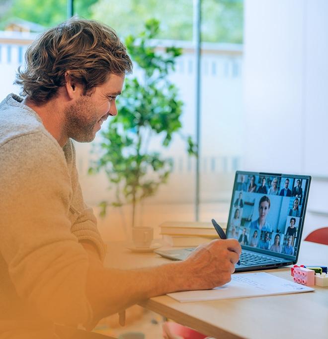 Studieninteressierter nimmt zuhause an einer Info-Veranstaltung teil. Er lächelt in den Laptop, wo die Teilnehmenden eines Online-Meetings zu sehen sind. Im Hintergrund steht eine Grünpflanze vor einem großen Fenster.