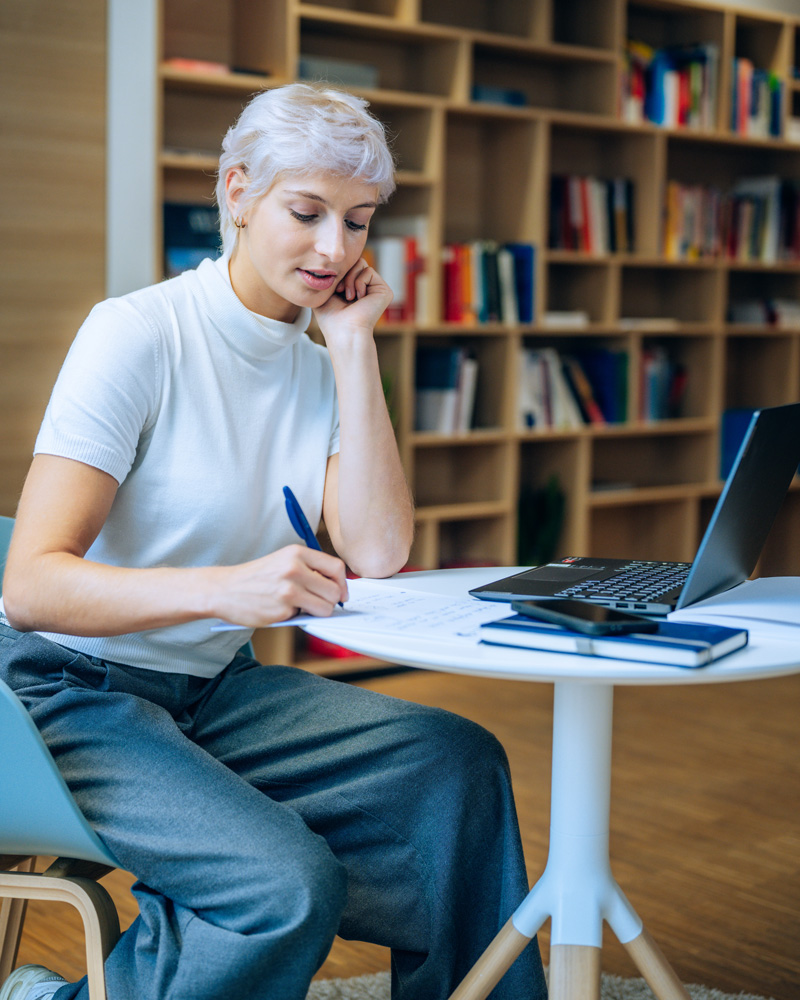 Student listens to an online lecture. She is taking notes. Next to her is a laptop at a small round table. A large bookshelf can be seen in the background.